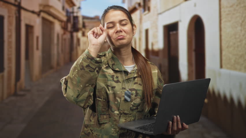 Woman soldier in camouflage holding laptop and rubbing her eye on a narrow sunlit street with dog tags visible; fatigue resilience determination.
