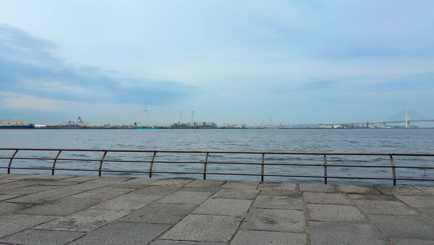 Wide shot of Yokohama port from Rinko Park in Japan. Cargo cranes and ships stand in the distant industrial harbor. Yokohama Bay Bridge is visible under a blue sky with soft clouds. Scenic waterfront view of the city bay area on a calm day.