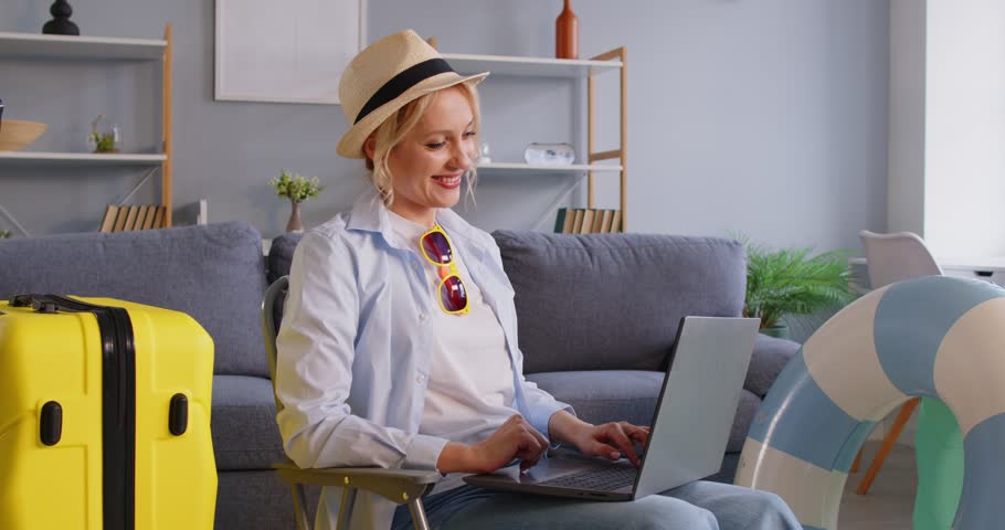 Happy excited young woman making yes gesture booking tickets online via laptop. Smiling girl in beach hat choosing tour for holiday trip sitting at home. Vacation concept. 4k video. Slow motion video