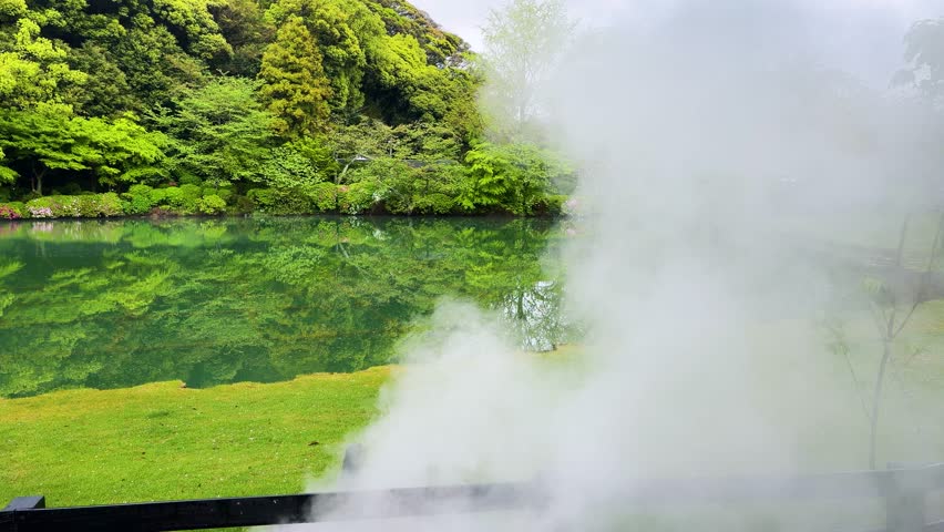 Beautiful Japanese garden pond surrounded by lush green trees and blooming bushes in Beppu. Heavy white steam rises from the thermal hot spring water surface. Calm mirror reflection of nature on the peaceful Shiraike Jigoku pond. Scenic geothermal landscape of traditional Japan park on a cloudy day.