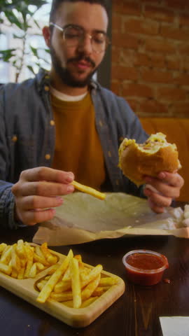 Vertical shot of man dipping fries into ketchup, sipping drink, holding burger while eating in restaurant