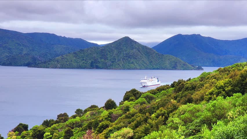 Blenheim, New Zealand - Ferry Sailing Through Marlborough Sounds Near Blenheim, New Zealand with Green Hills, Calm Water and Overcast Sky in a Scenic South Island Coastal View. The clip captures a scenic coastal landscape with a white ferry travelling across calm water surrounded by lush green hills and forested mountains. The overcast sky adds dramatic contrast to the vibrant landscape, highlighting the natural beauty of the Marlborough Sounds region. Ideal for travel, nature and coastal stock use showcasing South Island’s rugged terrain, ocean passages and peaceful maritime scenery.