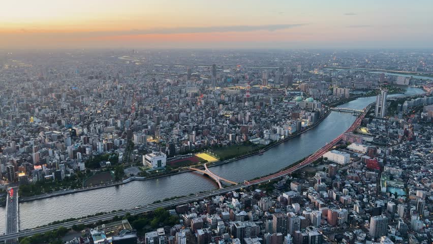 Aerial high-angle footage captures a panoramic evening view of Tokyo city from the Tokyo Skytree observation deck. The camera overlooks the winding Sumida River with its iconic Sakurabashi, Kototoibashi, and Shirahigebashi bridges. Residential and commercial skyscrapers spread across the vast urban landscape under a soft sunset sky. This static wide shot showcases the metropolitan skyline of Japan