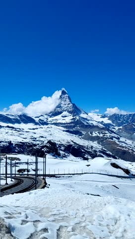 A stunning time-lapse video captures the iconic Matterhorn peak under a brilliant blue sky. The fast-motion footage highlights the dynamic movement of white clouds swirling around the sharp, pyramidal summit, while the snow-covered slopes of the Gornergrat region remain still below. In the foreground, the winding tracks of the Gornergrat Bahn cog railway lead the eye toward the majestic mountain range. The crisp alpine air and high-altitude sunlight emphasize the dramatic textures of the ice and rock. This vertical time-lapse showcases the interplay between the changing weather and the permanent grandeur of the Swiss Alps, offering a high-energy perspective on one of the world