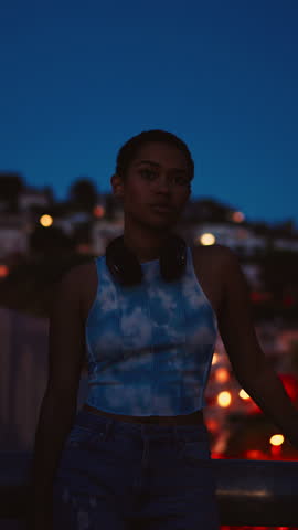 Young woman with headphones around her neck standing on balcony at twilight, gazing thoughtfully into the distance. Urban cityscape and soft evening glow create calm, contemplative, and stylish mood