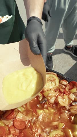 Vegetable stew and polenta are served onto a paper plate from a large pan during an outdoor food.