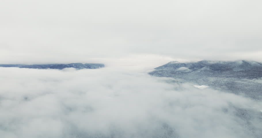 Drone flies left above a thick layer of winter clouds, revealing snow-covered Adirondack mountain peaks emerging through fog after fresh snowfall near Lake Placid, New York.