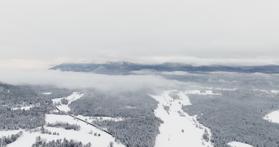 Drone fly in above the snow-covered Adirondack Mountains near Lake Placid, New York, as low winter clouds drift across ridgelines and fresh snowfall blankets forests and open fields below.