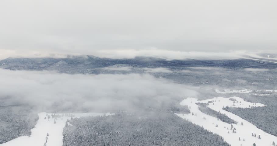 Drone flies above snow-covered open fields and forest as thick winter clouds drift across Adirondack mountain ridges near Lake Placid, New York, after fresh snowfall.
