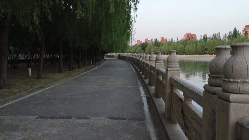 Cinematic tracking shot of a scenic riverside park path with lush greenery and calm water at sunset.