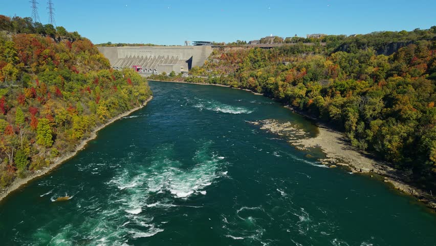 Powerful river flows past hydro-electric power station and autumn trees.