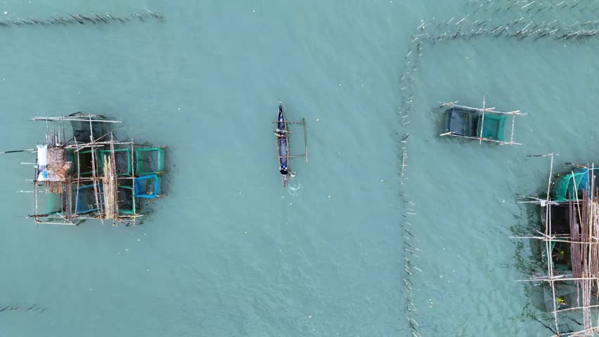 Aerial motion of fisherman with traditional boat casting fishing net over fish pens in blue sea.