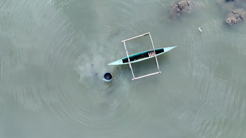 Top view of lone fisherman pulling nets near traditional boat in shallow tropical water during day.