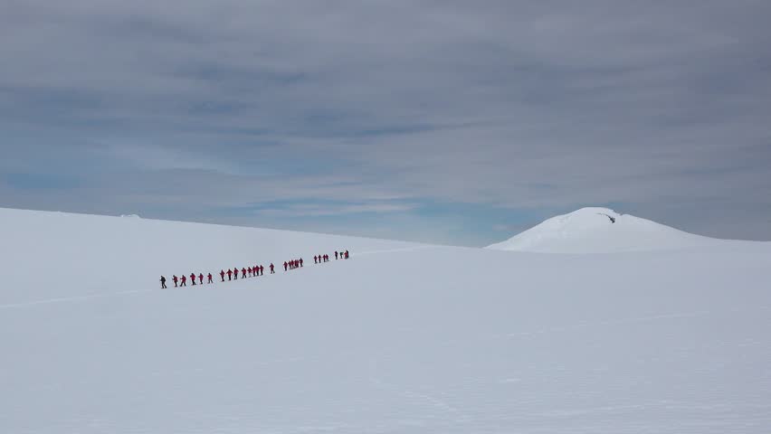 A group of people in bright jackets walk together over the vast white snow toward a tall mountain. The sky is gray and cloudy as they make their way through Antarctica.