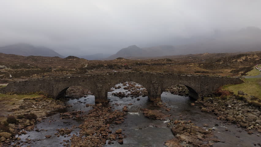 Panoramic ground shot of Sligachan Old Bridge, Isle of Skye. Cloudy sky reflects on the river below. Rugged Cuillin mountains in background. Scotland