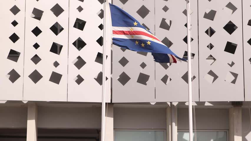 The national flag of Cabo Verde waving on a flagpole in front of a modern white building featuring a contemporary geometric diamond-patterned facade