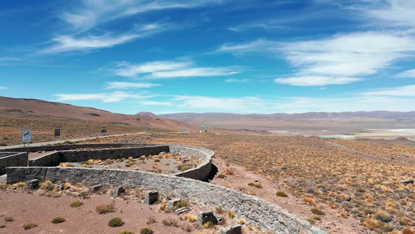 Forward flight over stone ruins of Paso Jama border checkpoint revealing vast arid plateau with salt lake and Andean mountain range under dramatic cloudy sky. Paso Jama, Chile