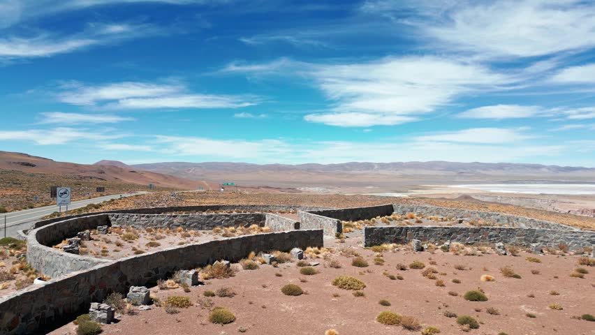 Panoramic aerial shot of abandoned stone border checkpoint ruins on high altitude plateau with salt lake and vast Andean mountain range under blue sky. Paso Jama, Chile