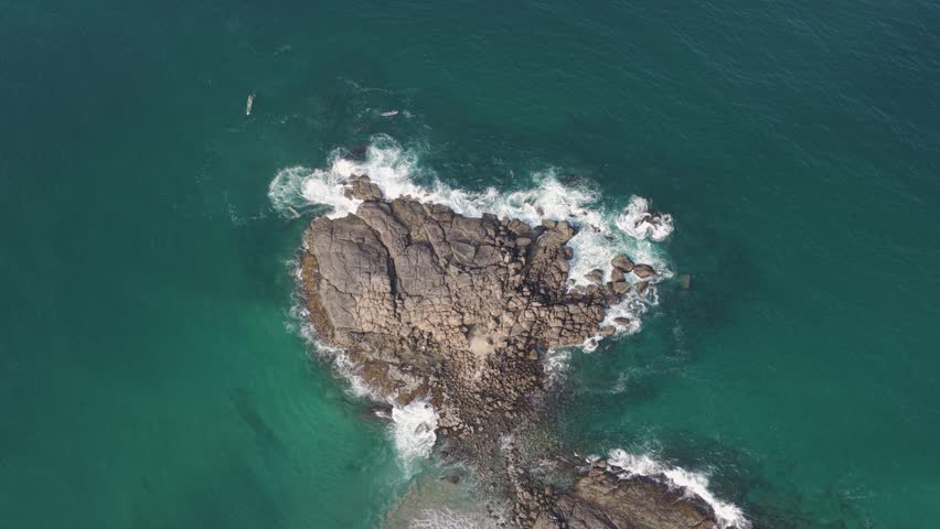 Aerial view of waves crashing against a rocky outcrop in the turquoise ocean with a small boat nearby in Kovalam, Kerala, India.