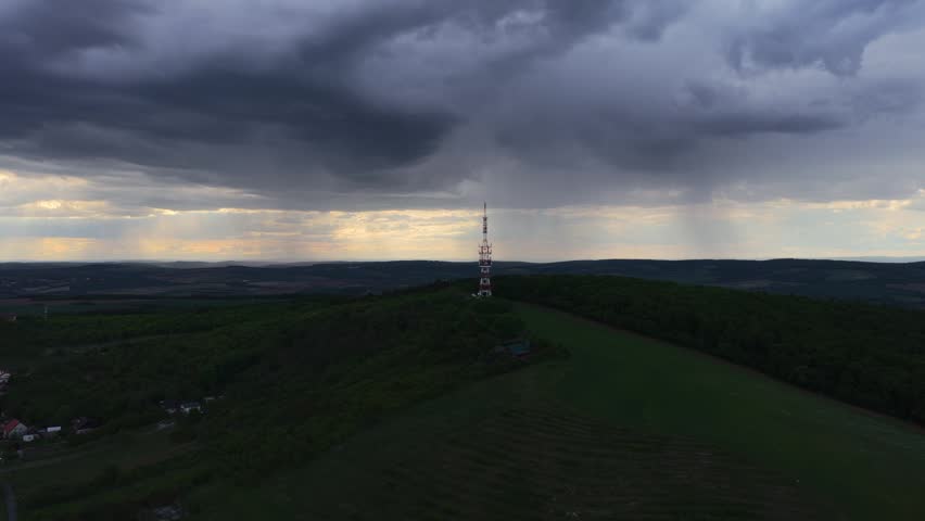 Aerial view of a communication tower on a green hill under dark storm clouds with rain falling in the distance Strazovice, South Moravian Region, Czechia.