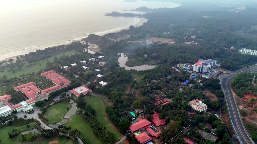 Cinematic aerial view of a tropical coastal landscape featuring luxury resorts, dense palm-covered greenery, winding water bodies, and a long sandy shoreline stretching beside the Arabian Sea.