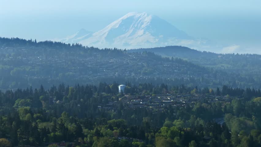 Bellevue Washington suburb district and lush green forest with massive snow-capped Mount Rainier in background under clear blue sky during sunny summer afternoon. Wide cinematic aerial shot.