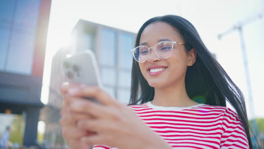 Happy woman wearing eyeglasses using smartphone near business center. Female smiling while browsing mobile application outdoors. Communicating online during sunny day. Wireless technology concept.