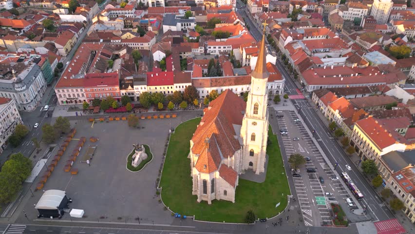 Aerial view of Cluj-Napoca city center with the Gothic-style St. Michael’s Church, its tall spire, red-tiled roofs, and dense historic buildings under warm sunset light