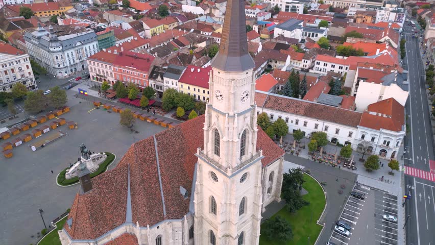 Aerial view of Cluj-Napoca city center with the Gothic-style St. Michael’s Church, its tall spire, red-tiled roofs, and dense historic buildings under warm sunset light