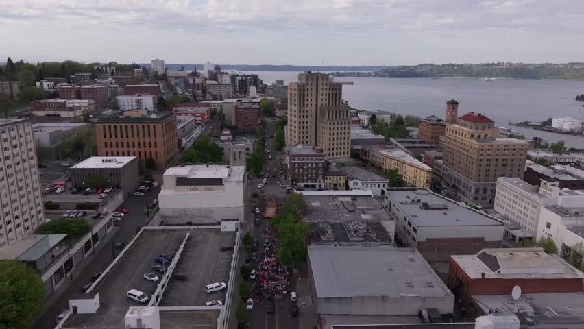 May Day Protest Crowd Marching On Street With View Of Old And New City Hall Of Tacoma On Commencement Bay. aerial pullback shot