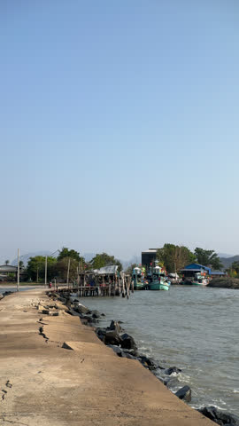 Sandy shoreline calm sea clear sky, empty beach with small harbor, moored boats near rocky jetty, distant buildings on low