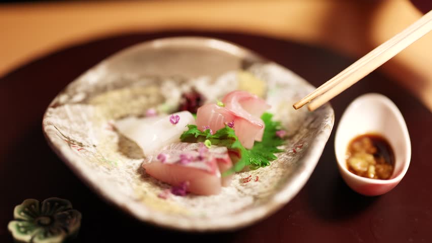 A close-up shot of chopsticks dipping raw fish into soy sauce on a ceramic plate