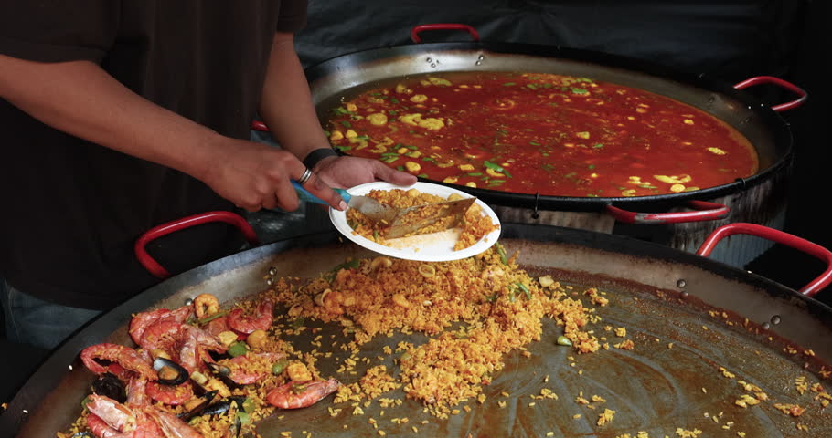 Street food vendor serves fresh seafood paella from large traditional metal pan into white plate. Chef hands use metal spatula to scoop yellow rice with mussels shrimps and vegetables during outdoor