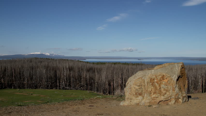 Scenic overlook with large rock and Yellowstone Lake in background. Snowcapped mountains rise in the distance beyond a landscape shaped by past wildfires. A wide natural vista highlighting the dramatic beauty of the American West.