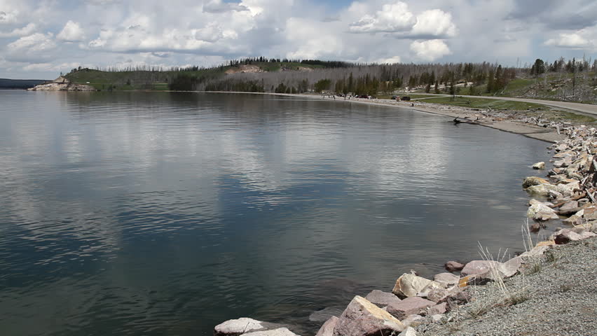 Scenic lakeside road following the rocky shoreline of Yellowstone Lake. Green hills and forested slopes stretch toward distant mountains under dramatic clouds. A wide travel landscape capturing the expansive, open character of Yellowstone’s protected environment.