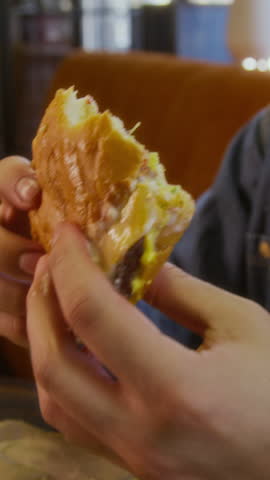 Vertical close up shot of biracial man holding cheeseburger, taking bite, enjoying meal at restaurant table