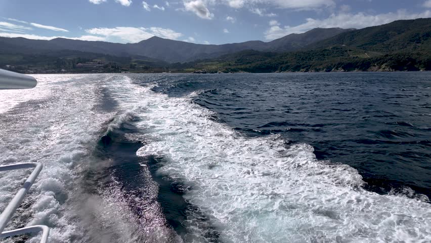 The boat moves through water, creating waves behind it. Mountains stand tall in the distance under a clear sky. The sun shines on the water during day hours.