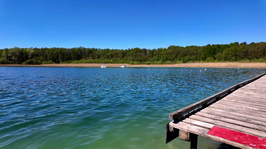 Sunny lake landscape featuring a wooden pier in the foreground, crystal clear turquoise water with visible sandy bottom in the shallows, lush green forest background under a bright blue cloudless sky.