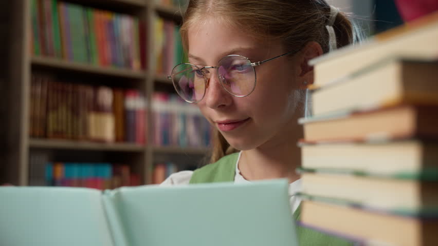 Happy clever Caucasian little girl child school kid in eyeglasses reading book turning pages looking at camera daughter smiling elementary studying in library pupil wearing glasses read study smile