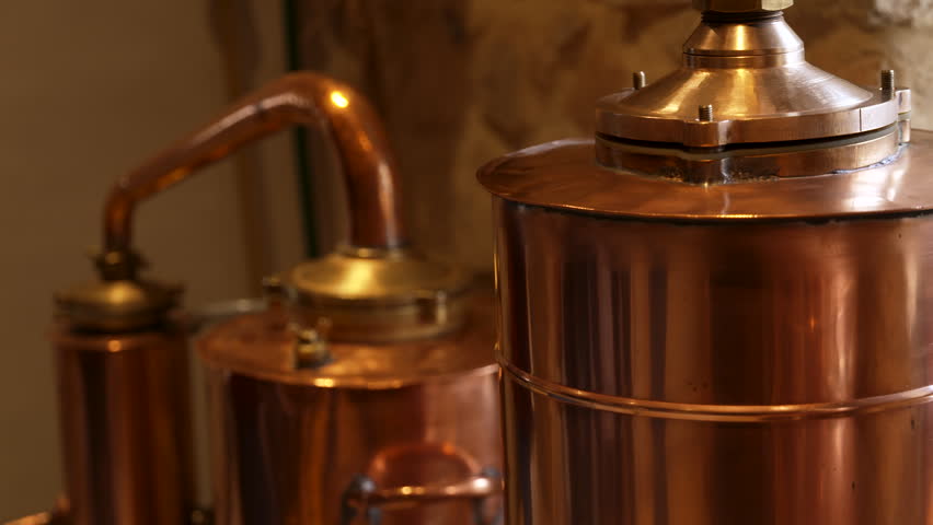 Close-up of a traditional copper absinthe still inside a distillery, with warm ambient light reflecting on the metal surfaces. Static shot highlighting artisanal distillation equipment, craftsmanship, and the authentic atmosphere of spirit production.