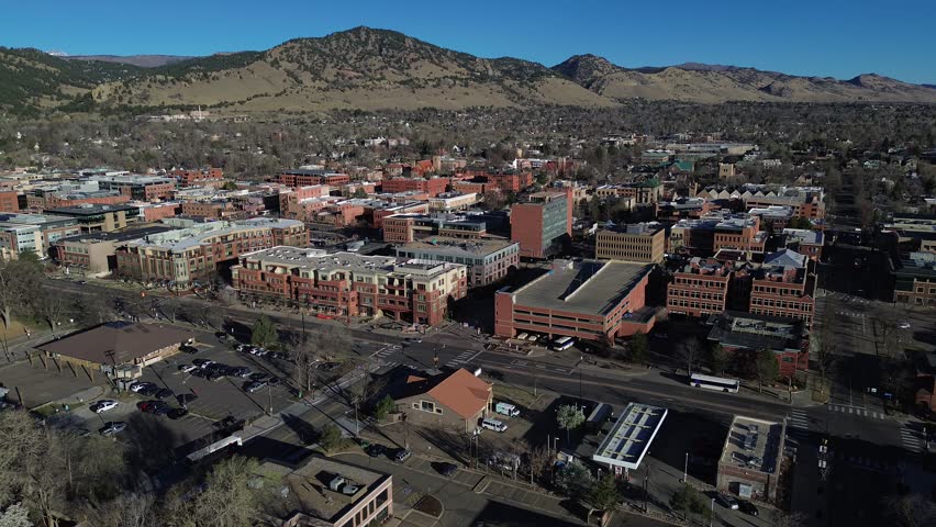 Creek adjacent downtown blocks overlook Boulder civic corridor near Canyon Blvd, commercial roofs, parking courts. Varied surfaces, muted foliage, clear directional light urban foreground, CO. USA