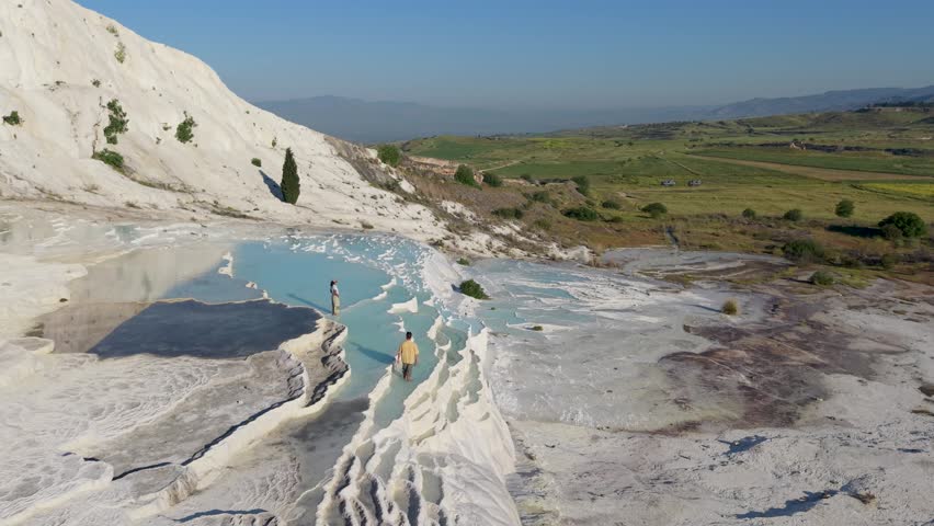 A couple explores the mesmerizing travertine terraces of Pamukkale, Turkey, where turquoise waters cascade over white mineral-rich formations. Experience the beauty of this natural wonder.