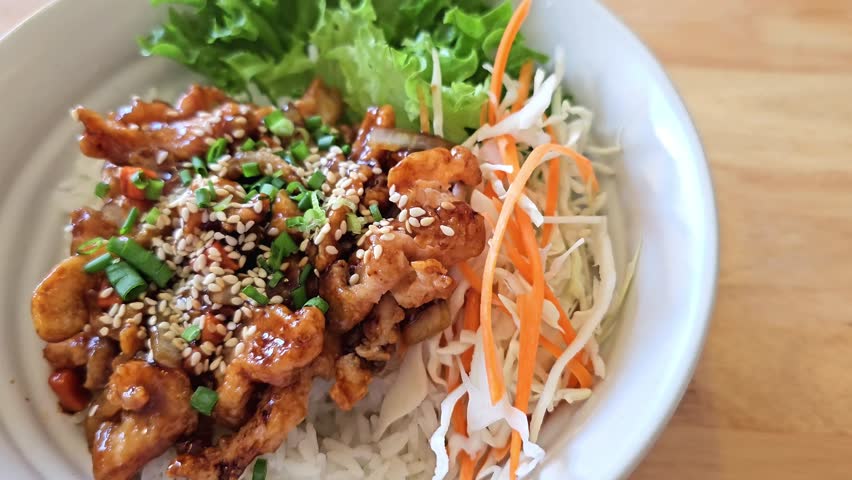 Asian rice bowl with fried chicken, sesame seeds, green onion, cabbage salad and lettuce in ceramic bowl on wooden table. Traditional homemade meal with vegetables and sauce. Close up shot with selective focus