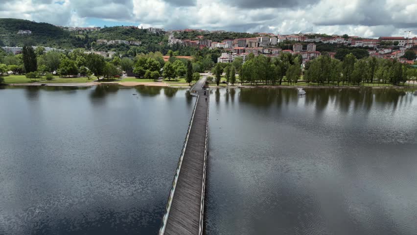 Aerial shot of a wooden pedestrian footbridge with colorful geometric railings crossing a calm section of the Mondego River at a green riverside park with people walking and kayaking