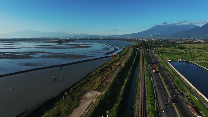Clear skies above a river and highway. Vehicles move along a wide asphalt road. Mountains loom in the distance, serene and tall. Green grass borders the riverbanks and the road. No people are visible