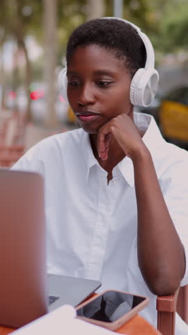 Happy and engaged young Black woman participating in work meeting or webinar, wearing headphones and using laptop computer at outdoor café. Female freelancer enjoying online video call on netbook