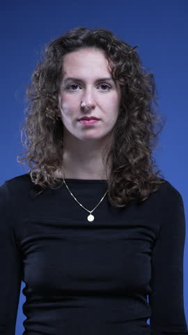 Portrait of young woman with curly hair looking at camera with neutral expression on blue background, vertical video