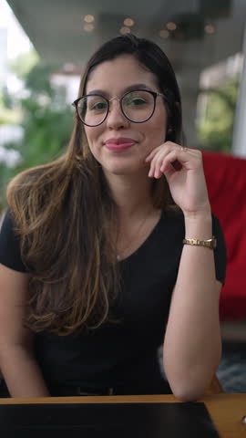 One happy young woman smiling at camera. Charming 20s girl wearing eyeglasses seated at coffee shop in Vertical Video