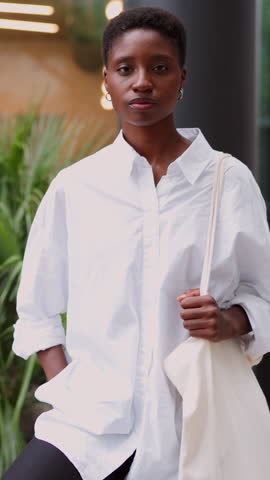 portrait of determined and confident young Black woman standing in modern urban setting posing proudly. Stylish female wearing white shirt and carrying a tote bag, exuding strength and resolve