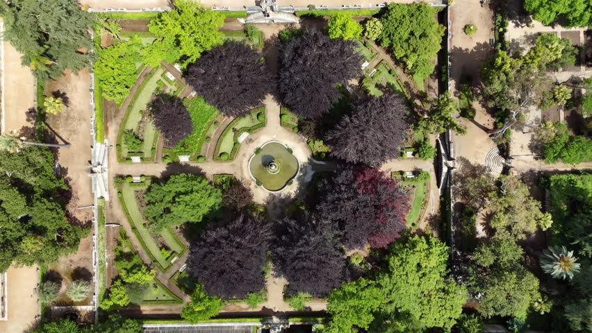 Overhead aerial view of a historic botanical garden with symmetrical pathways central fountain purple and green trees and terraced landscaping seen from directly above